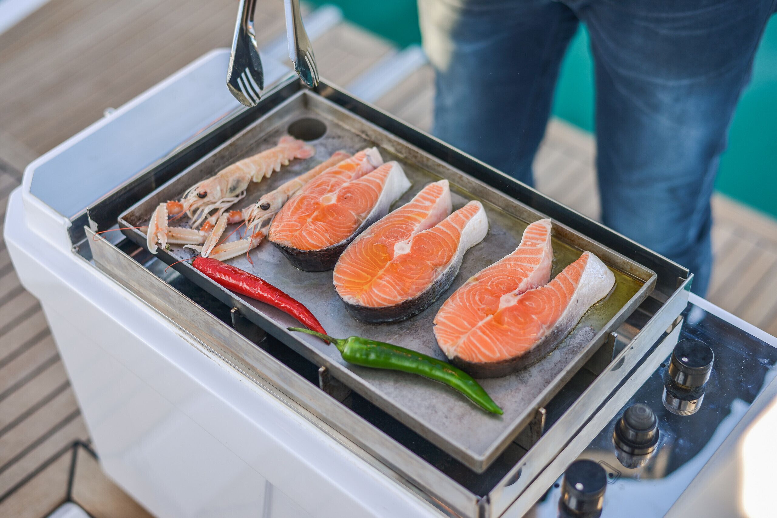 Ein tragbarer Grill mit Lachs und Garnelen auf einer Grillmatte, eine rote Chili-Schote und eine grüne Chili-Schote.