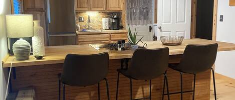 Una cocina con armarios de madera, un refrigerador, una cafetera y un comedor con taburetes de bar.