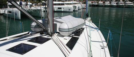 Un port de plaisance où sont amarrés de nombreux bateaux, un voilier à la coque blanche et au pont fermé, et un paysage montagneux en arrière-plan.