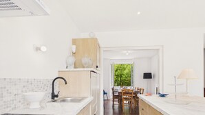 Una cocina moderna con un lavamanos, estufa y armarios de madera. Se observa un área de comedor con una mesa y sillas a través de un vano abierto.