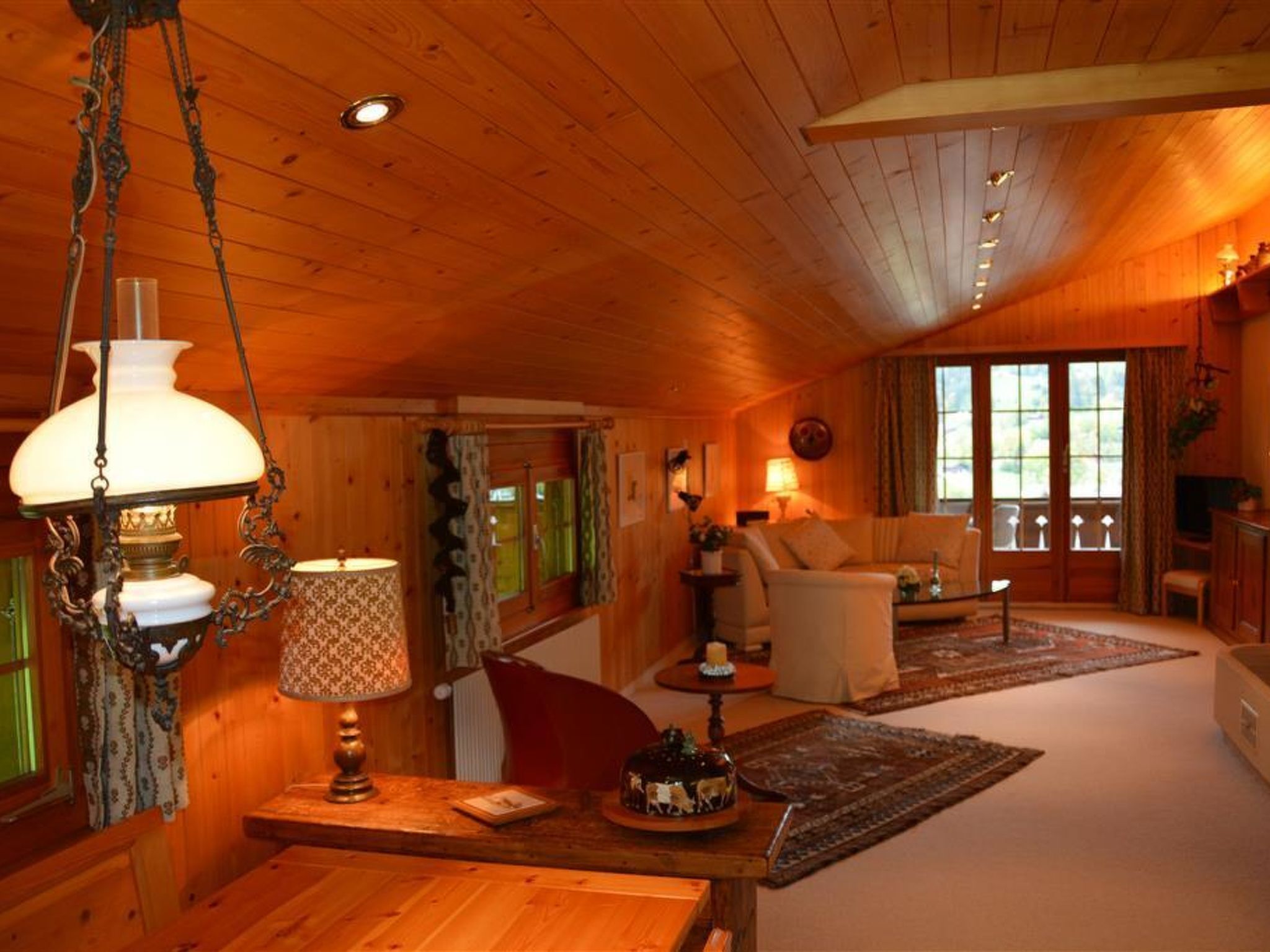 A cozy cabin interior with wooden walls, a dining area, a living room with a sofa and armchairs, and a large window providing natural light.