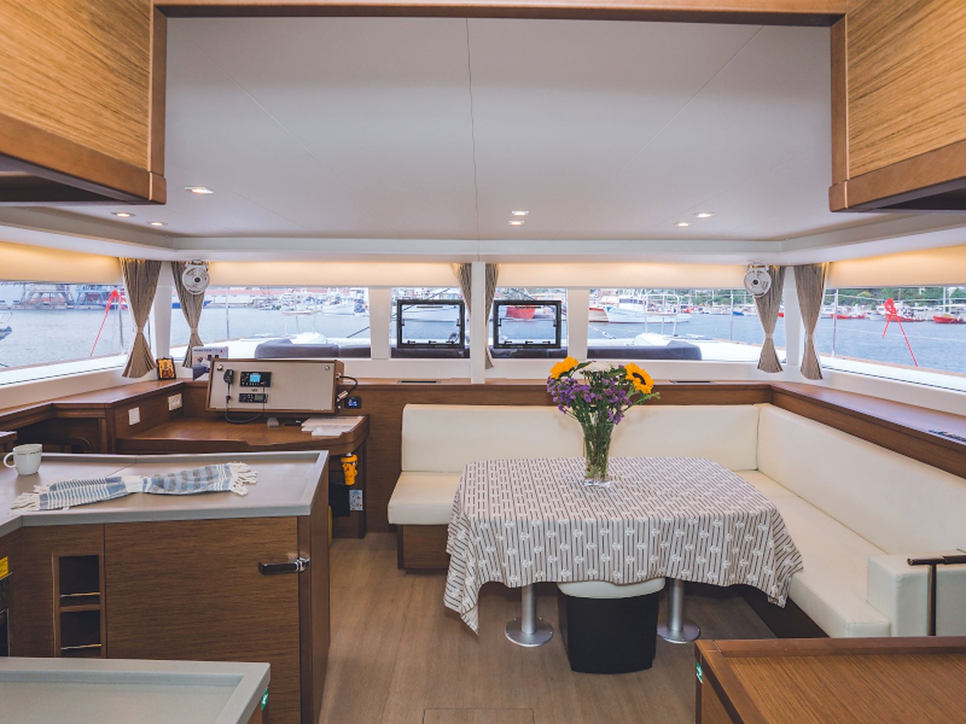 Interior de barco com área de refeições, painel de controle e vista para a marina.