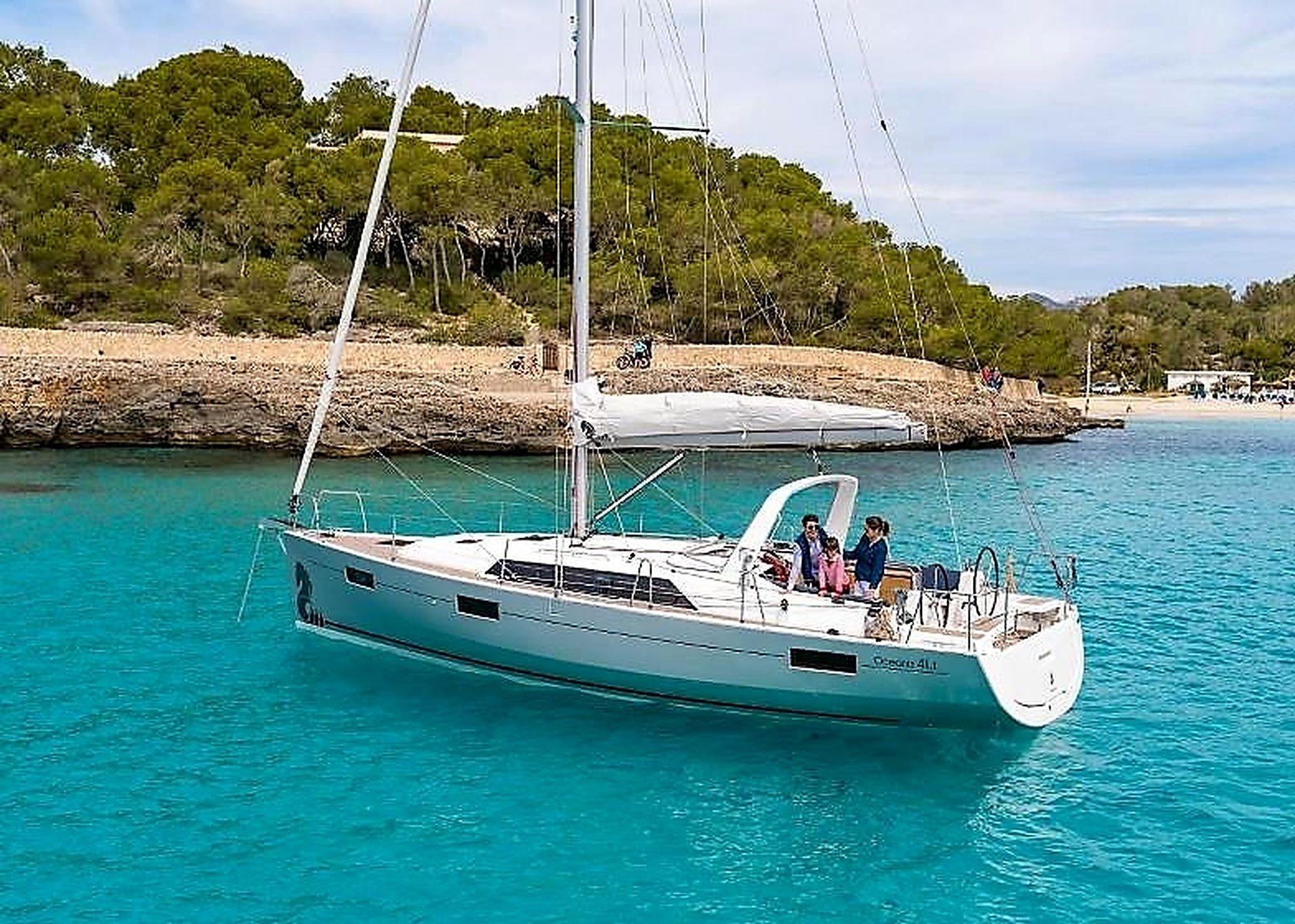 Eine Segelyacht mit Kabine und überdachter Deck, vertäut in der Nähe einer felsigen Küste, im Hintergrund Bäume.