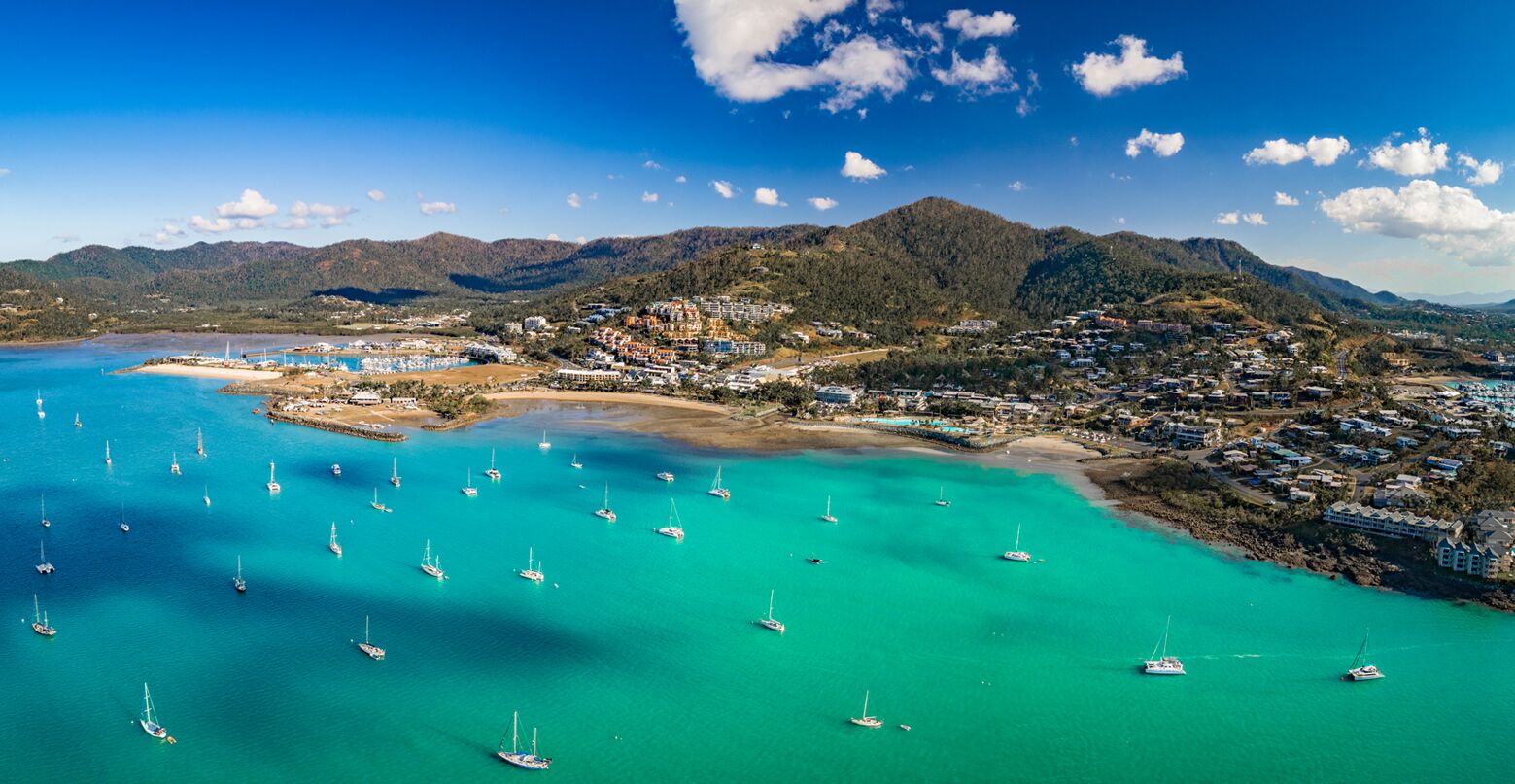A coastal town with clear turquoise waters, numerous sailboats, and a mountainous backdrop.
