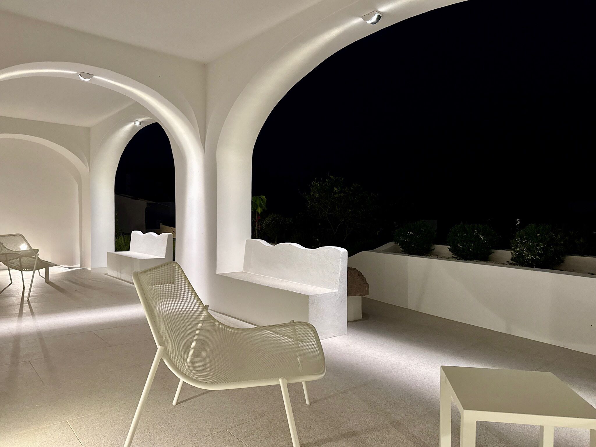 Un espacio de estar al aire libre con sillas y mesas blancas, bajo un techo arqueado blanco, de noche.