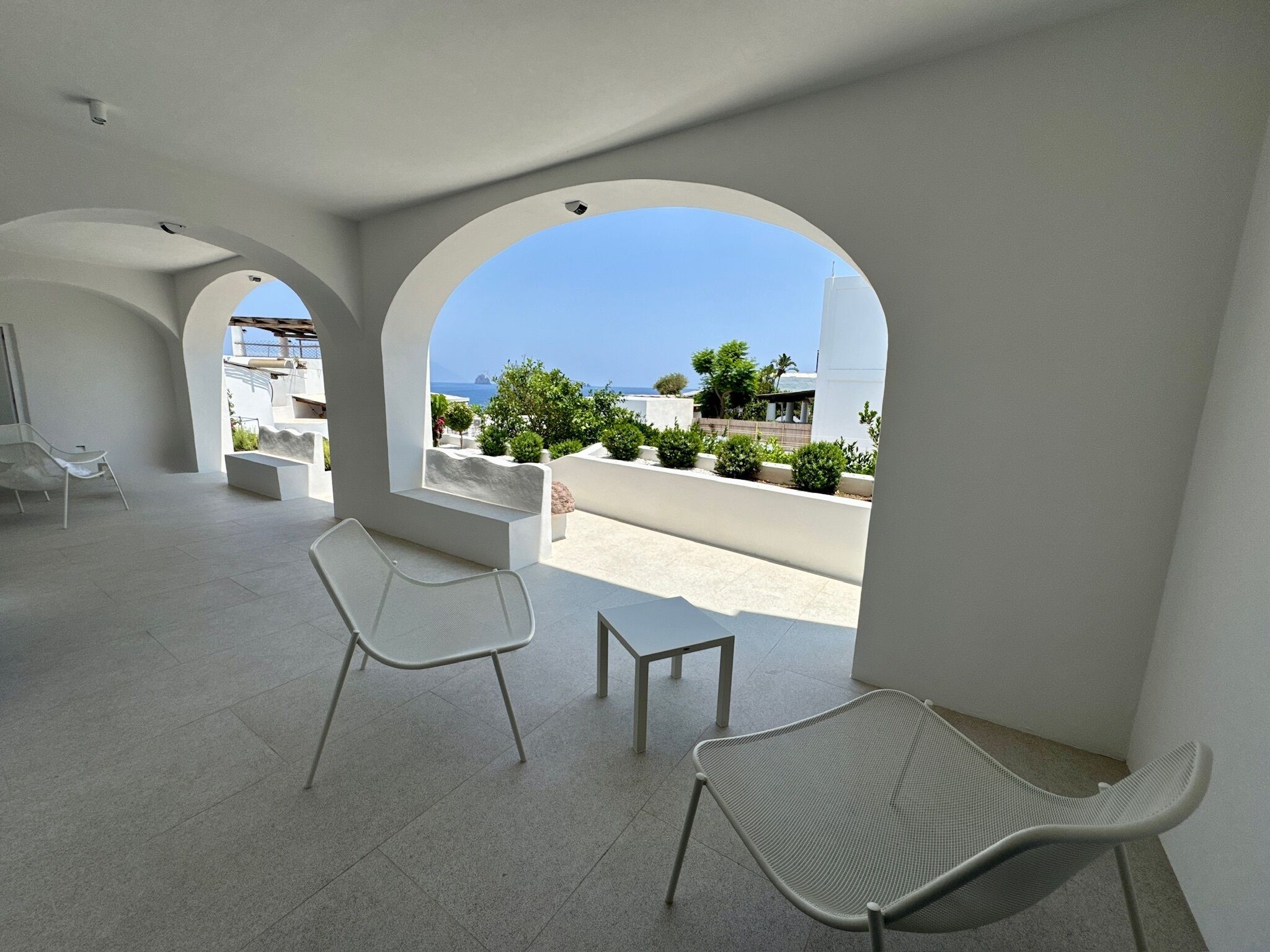 A modern interior with white walls and arches, a seating area with chairs, and a view of the sea.