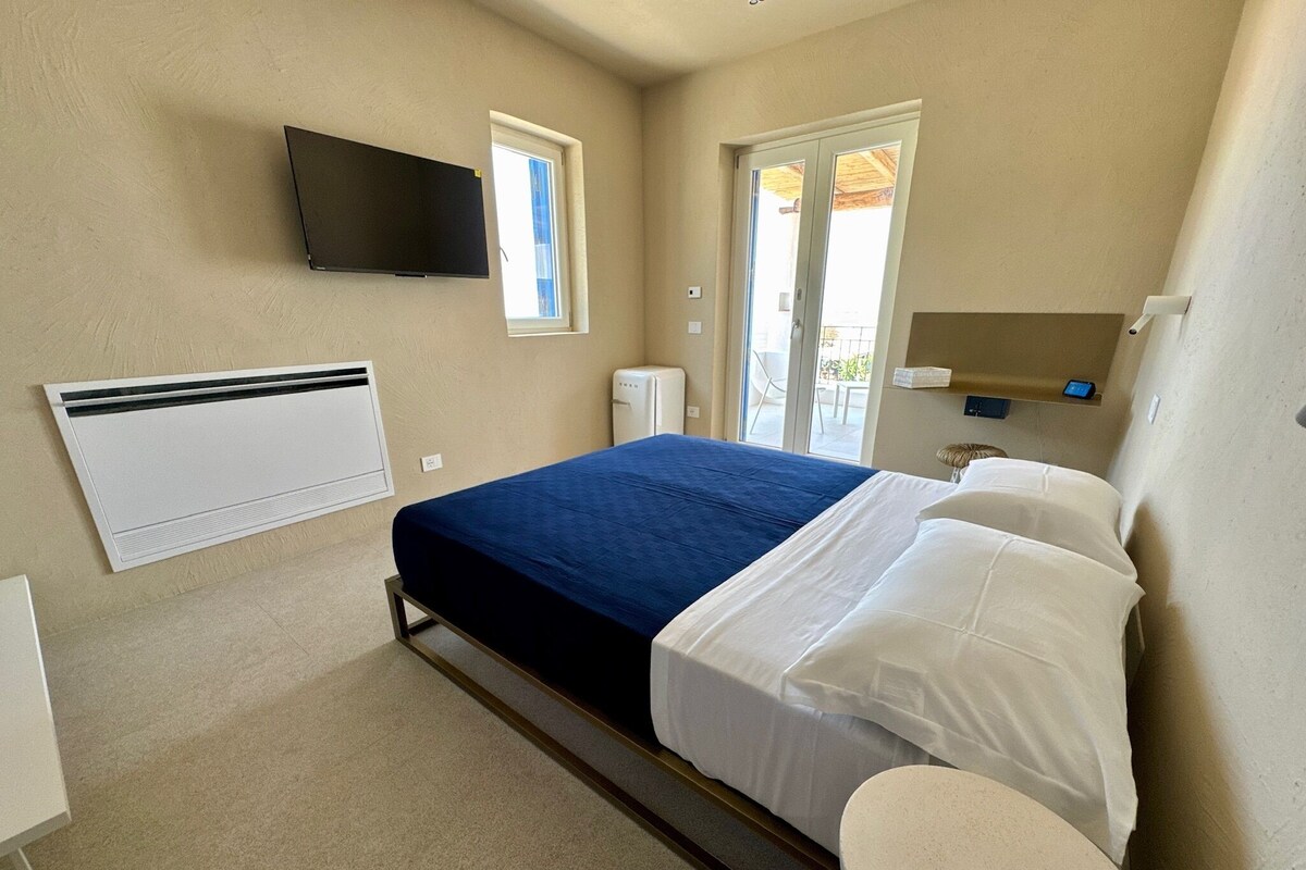 A bedroom with a bed, a television, a heater, a refrigerator, and a toilet.