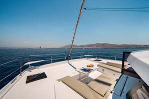 A boat deck with seating, a table with a fruit bowl, and a view of the sea and distant land. - Sun-Kissed Sailing Adventure in the Ionian Islands (Nikiana)