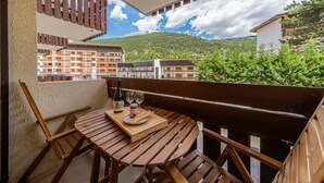 Un balcone con un tavolo di legno, una bottiglia e dei bicchieri, affacciato su un paesaggio montuoso con edifici.