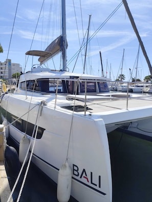 Un yate blanco llamado 'BALI' amarrado en un puerto deportivo con otras embarcaciones, cielos despejados y edificios al fondo.