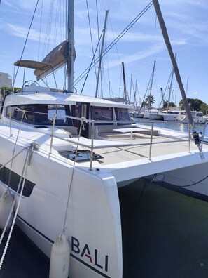 A white yacht named 'BALI' docked at a marina with other boats, clear skies, and a calm sea.