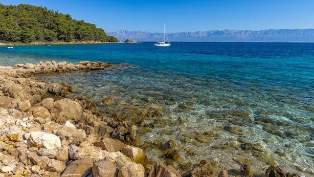 Una costa rocciosa con acque limpide, una barca a vela e una collina boscosa sullo sfondo.