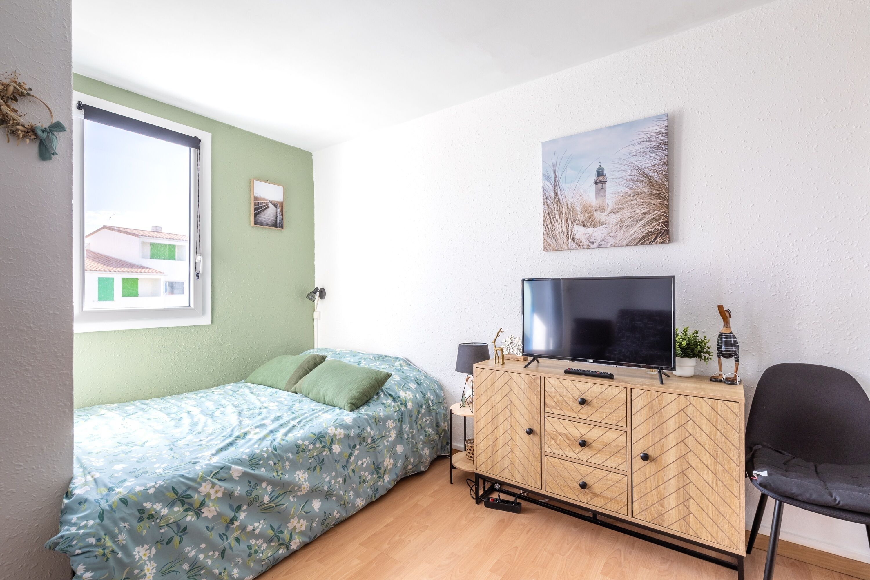 Ein Schlafzimmer mit einem Bett, einem Fernseher, einem Holzschrank, einem Sessel und einem Fenster mit Blick auf Gebäude.