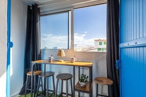 Ein Eckzimmer mit Fenster, einem Tisch mit Hockern, einer Topfpflanze und einer blauen Tür.