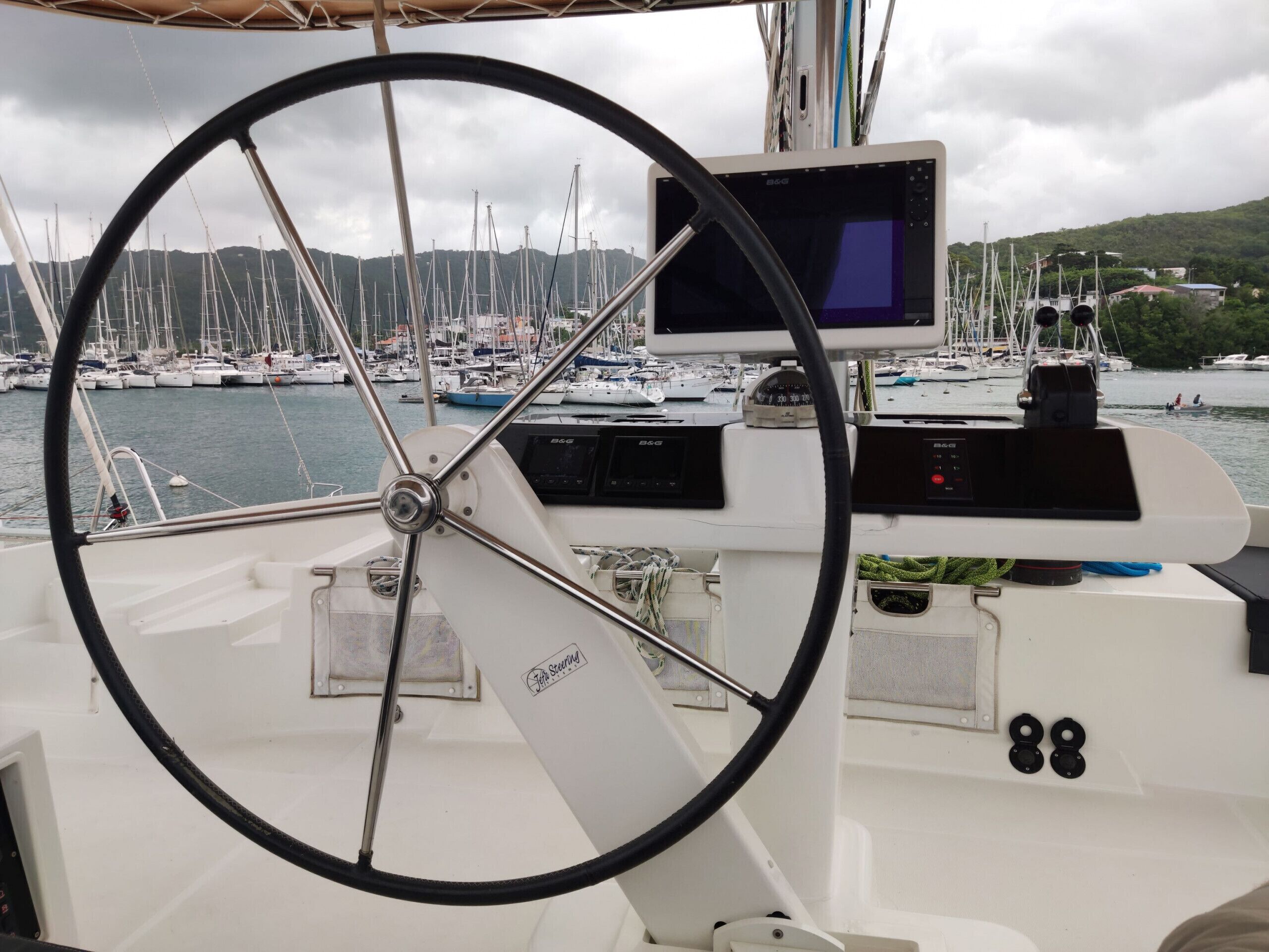 Timón de barco, panel de control y un puerto deportivo con varias embarcaciones al fondo.