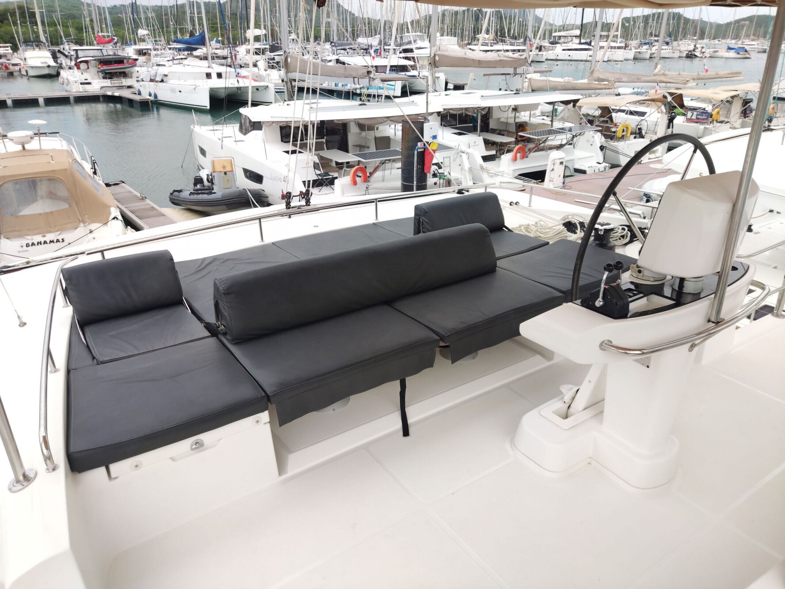 Un yacht doté d’un espace salon avec des coussins noirs, d’un volant et d’une vue sur une marina où sont amarrés de nombreux bateaux.