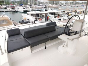 Un yacht doté d’un espace salon avec des coussins noirs, d’un volant et d’une vue sur une marina où sont amarrés de nombreux bateaux.