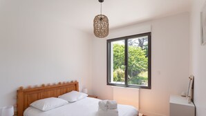 Een slaapkamer met een houten bed, witte beddengoed, een nachtkastje met een lamp, een raam met uitzicht op bomen, en een hangende lamp.