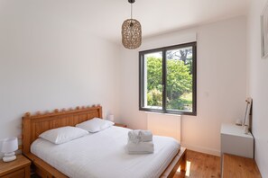 A bedroom with a wooden bed, white bedding, a bedside table with a lamp, a window with a view of trees, and a hanging light fixture. - Jardin papillons - À moins d'1 km de la plage (Erquy)
