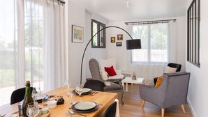 Een eetruimte met een houten tafel, stoelen en een fles wijn. Er zijn ramen met gordijnen, een lamp en lijstjes met afbeeldingen aan de muur.