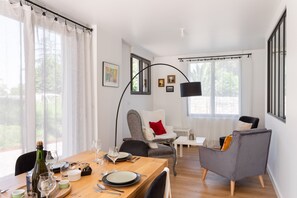 A dining area with a wooden table, chairs, and a bottle of wine. There are windows with curtains, a lamp, and framed pictures on the wall. - Jardin papillons - À moins d'1 km de la plage (Erquy)