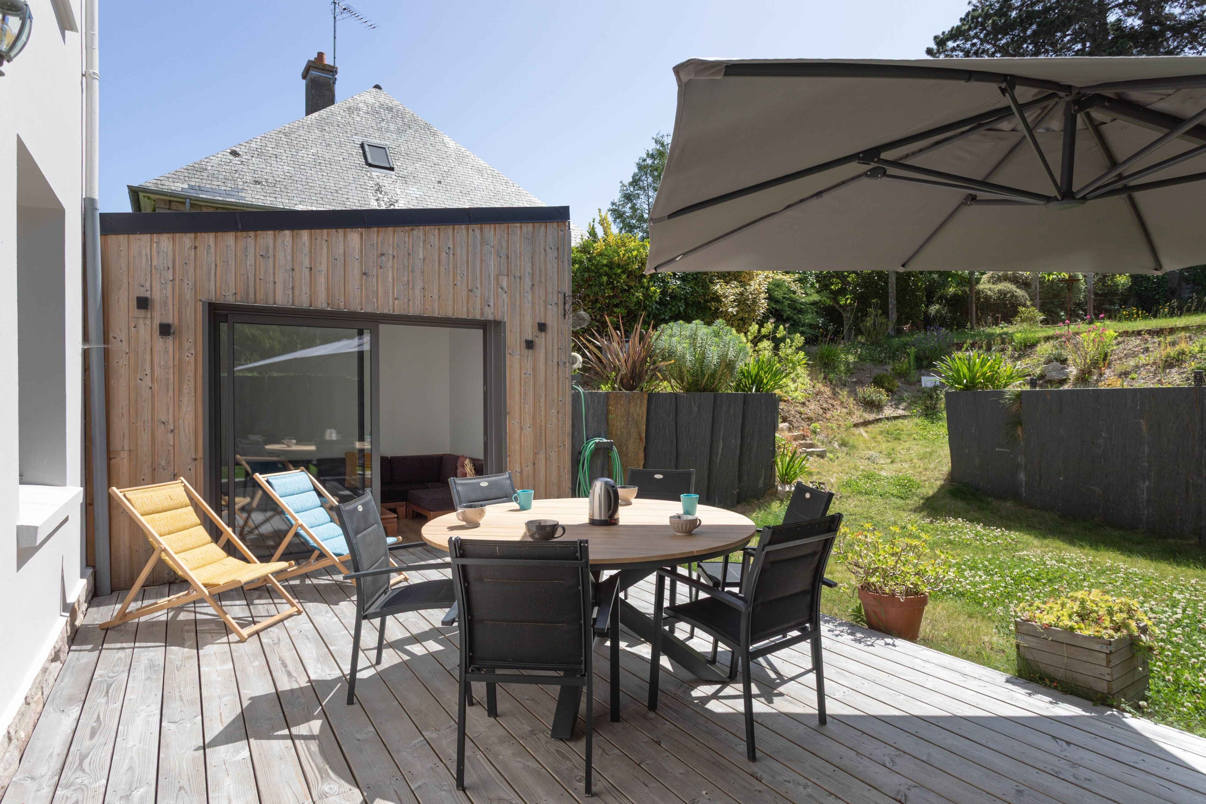 Eine Terrasse im Freien mit Holzdeck, einem runden Tisch mit Stühlen, einem großen Sonnenschirm und einem Haus im Hintergrund.