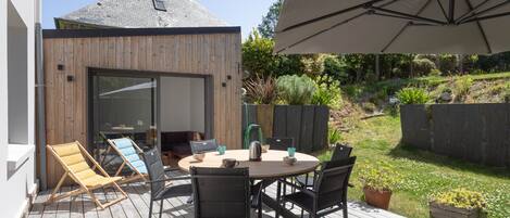 Eine Terrasse im Freien mit Holzdeck, einem runden Tisch mit Stühlen, einem großen Sonnenschirm und einem Haus im Hintergrund.