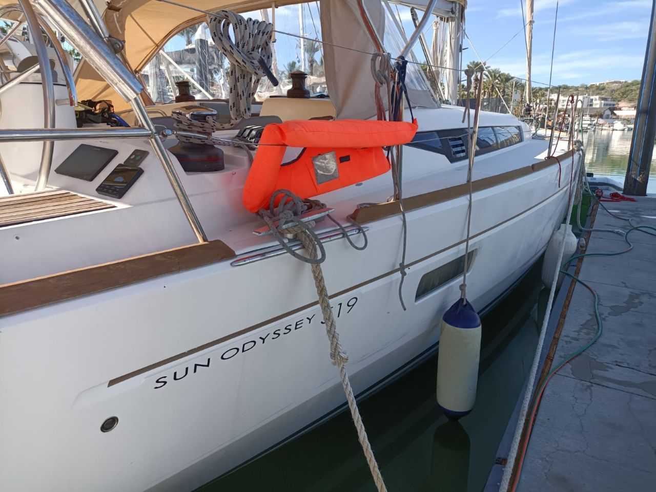 Eine weiße Yacht namens SUN ODYSSEY 19 liegt in einer Marina festgemacht, mit Festzurrseilen, einer Rettungsboje und einer Armaturenkonsole.