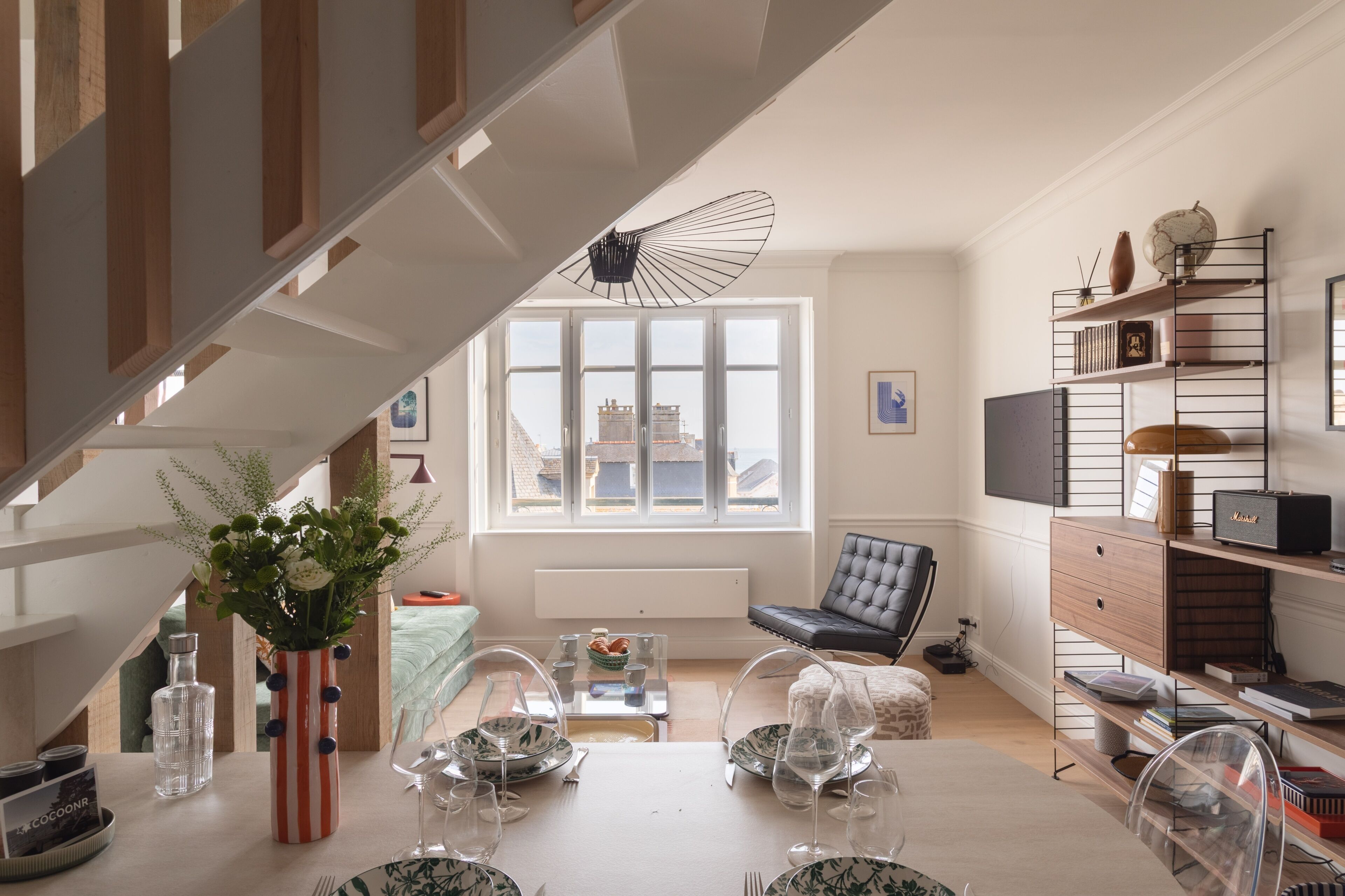 Ein modernes Wohnzimmer mit Essbereich, einer Treppe und Blick auf die Stadt durch große Fenster.