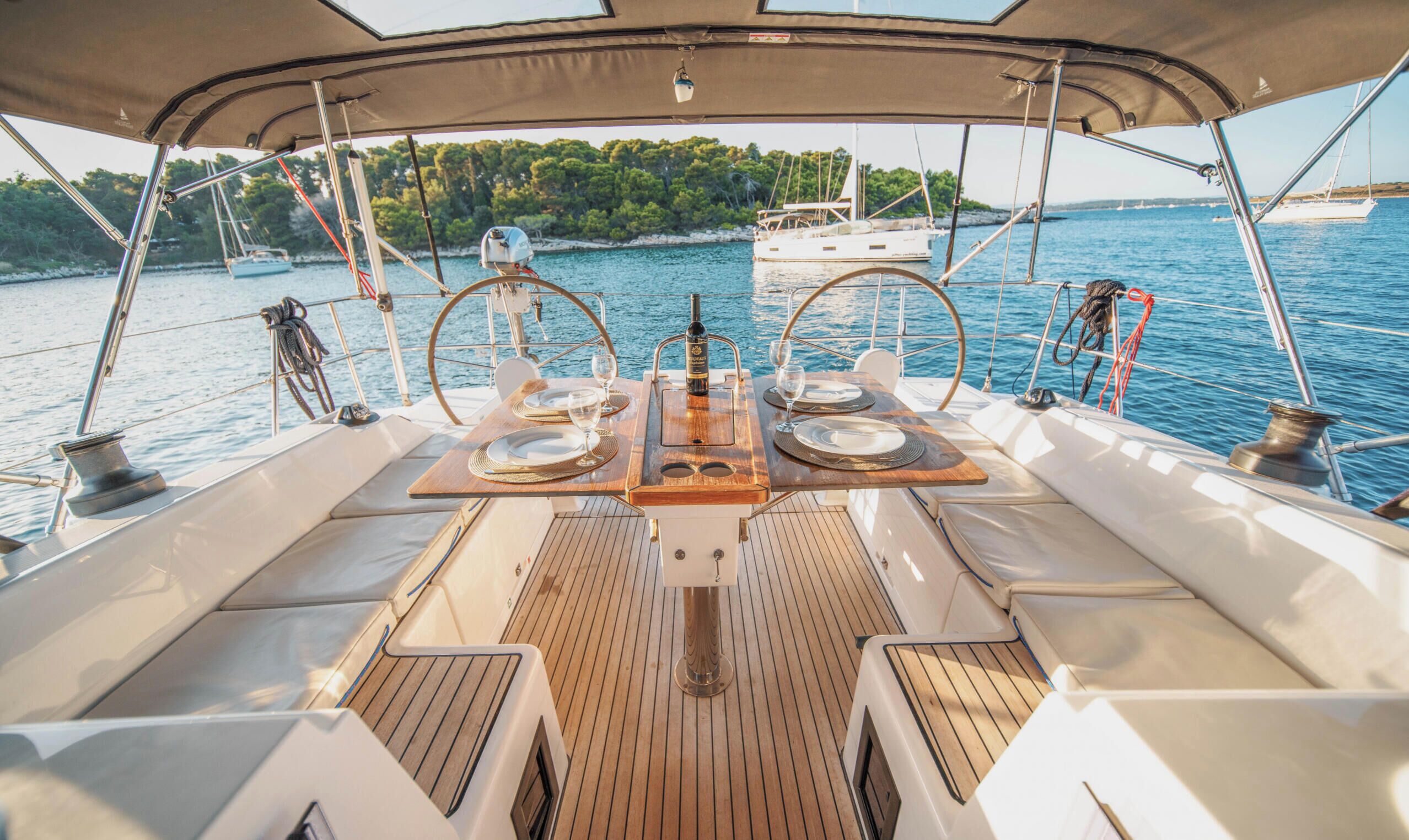 A boat deck with a table set for two, a bottle of wine, and a view of the sea and trees.