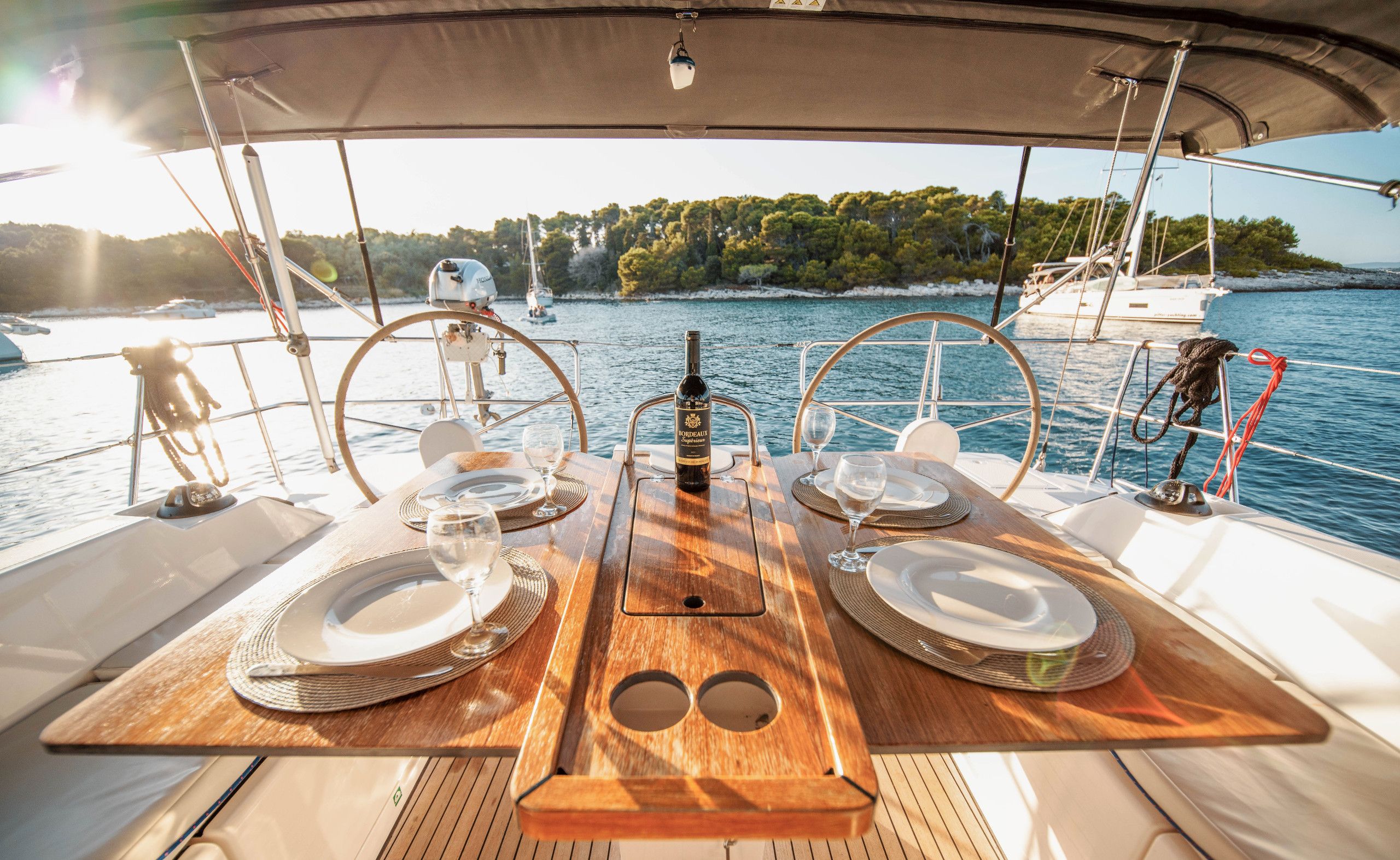 A dining table set for two on a boat with a view of the sea and a forested coastline.