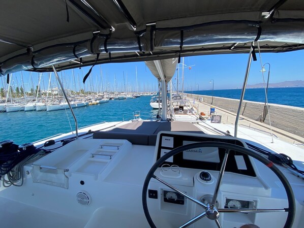 A boat's interior with a steering wheel, control panel, and a view of the marina.