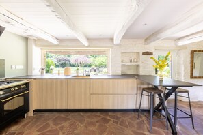 A kitchen with a stone floor, wooden cabinets, a black stove, and a large window with a view of greenery. - La Chaumière BZH - Pour 5 avec grand jardin (Riec-sur-Bélon)