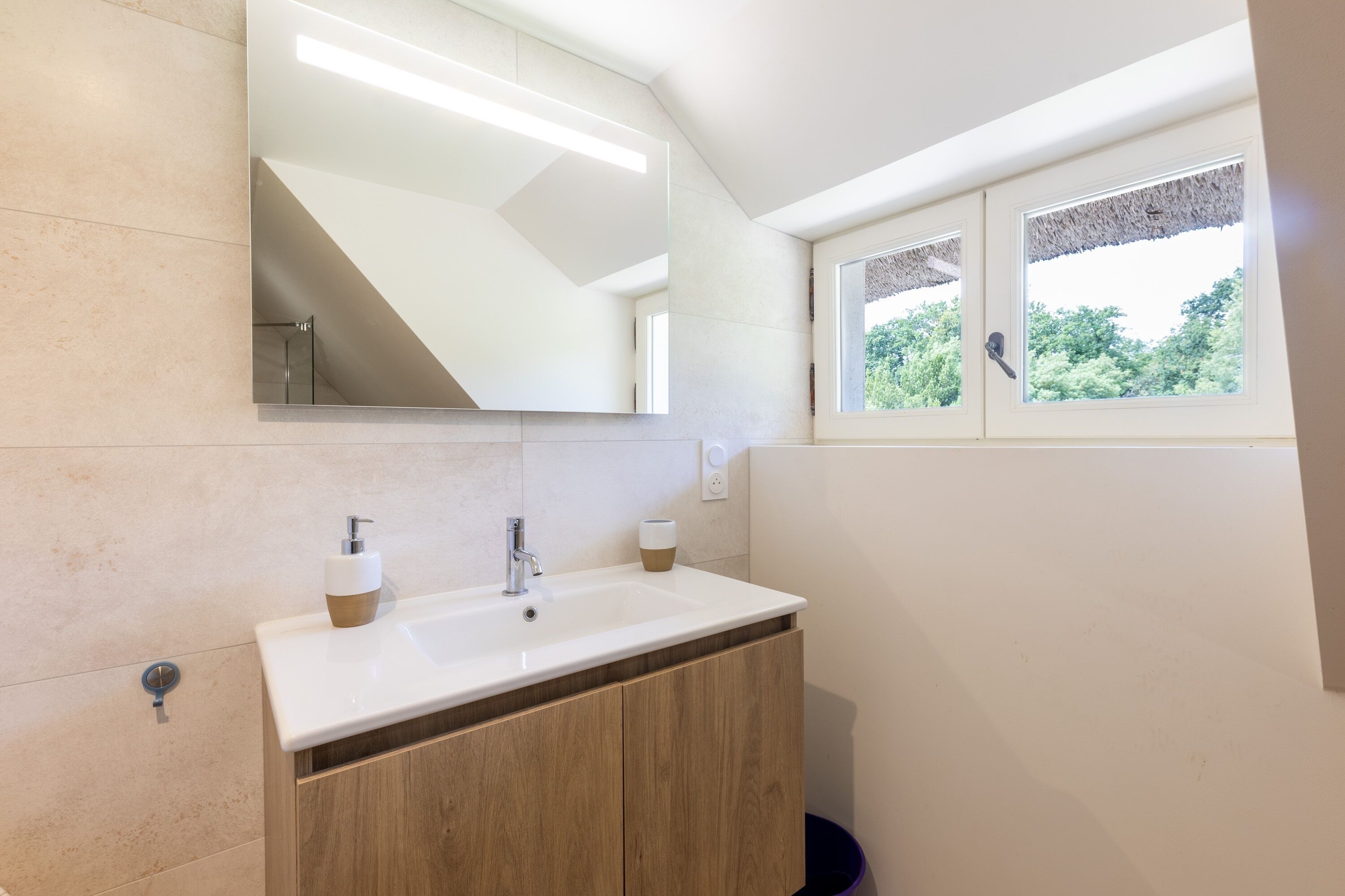 A bathroom with a white sink, wooden cabinets, a mirror, and a window with a view of greenery.