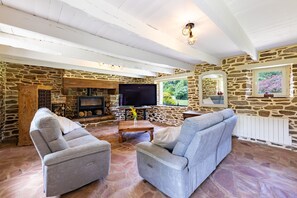 A stone-walled living room with a fireplace, a television, and two grey sofas. - La Chaumière BZH - Pour 5 avec grand jardin (Riec-sur-Bélon)