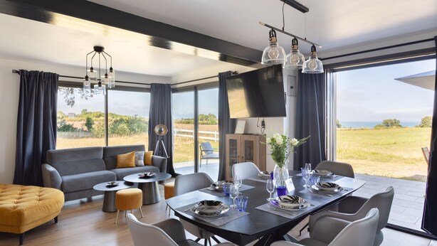 A modern dining area with a table set for four, a sofa, and a large window with a view of the outdoors. - La Corniche - Belle maison pour 6 avec vue mer (Cancale)