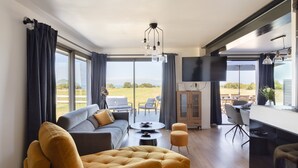 A modern living room with a sofa, ottoman, and dining area, large windows with curtains, and a flat-screen TV. - La Corniche - Belle maison pour 6 avec vue mer (Cancale)
