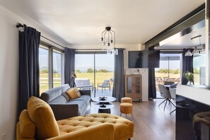 A modern living room with a sofa, ottoman, and dining area, large windows with curtains, and a flat-screen TV. - La Corniche - Belle maison pour 6 avec vue mer (Cancale)