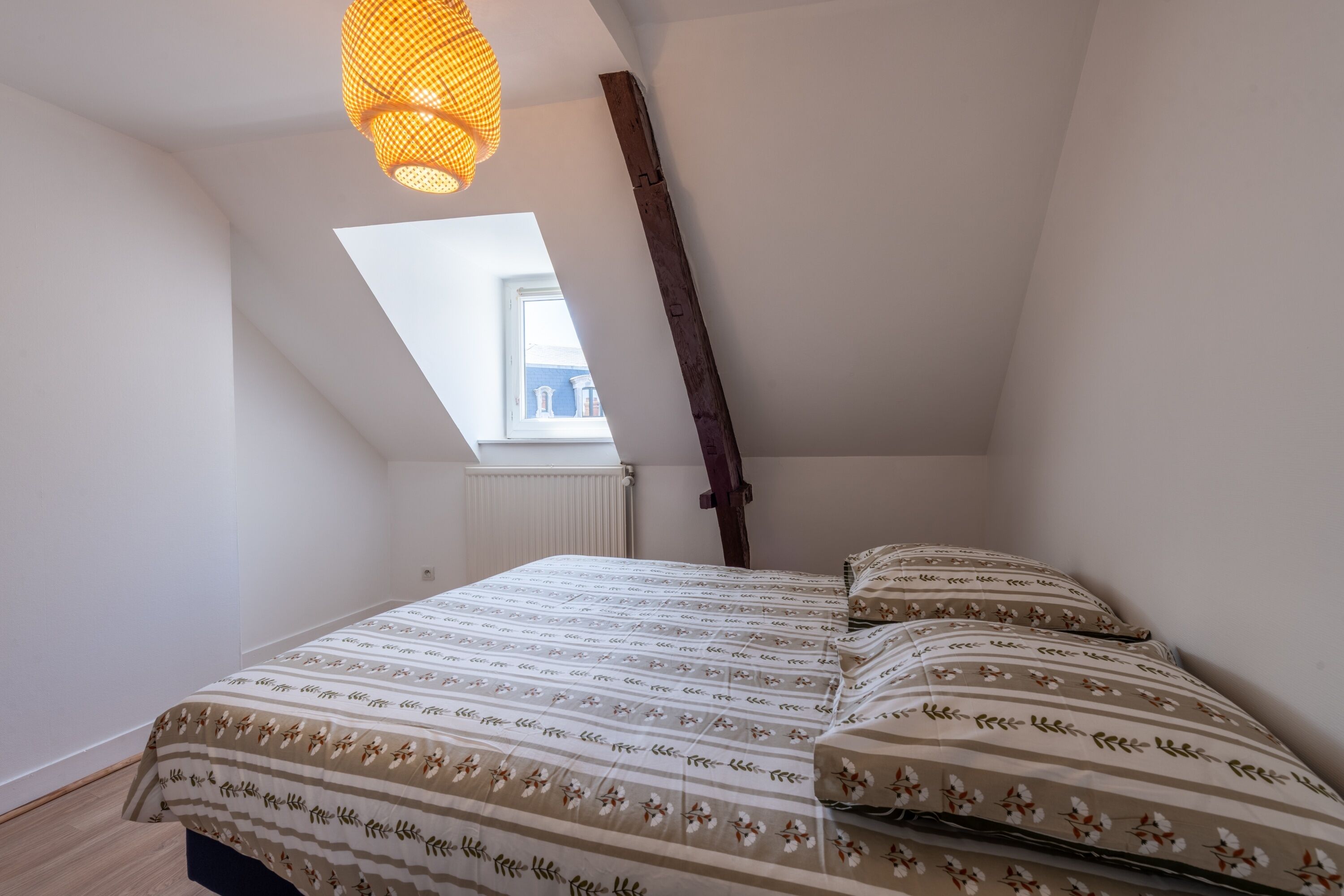 Ett sovrum med ett sluttande tak, en säng med mönstrat sänglinne, en hängande lampa och ett fönster med utsikt över byggnader.