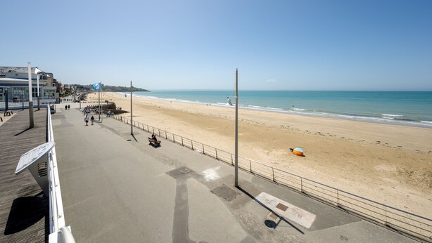 Blick auf einen Strand mit Promenadenweg, Strandsonnenschirmen und blauem Himmel.