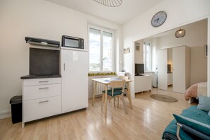 A compact living space with a kitchenette, a dining area, a bed, and a view through a large window. - Le Studio du Val - 2min à pied de la plage (Pléneuf-Val-André)