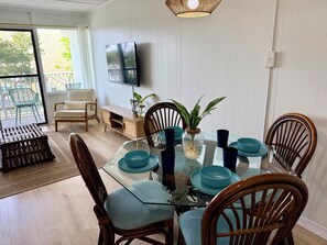 Een eetgedeelte met een glazen tafel, blauwe stoelen en een houten kast met een televisie aan de muur.
