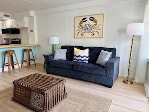 A living room with a dark blue sofa, two wooden stools, a coffee table, a lamp, and a framed artwork on the wall. - Oceanside Condo - Sleeps 6 - Beach Gear - Parking (Ocean City)