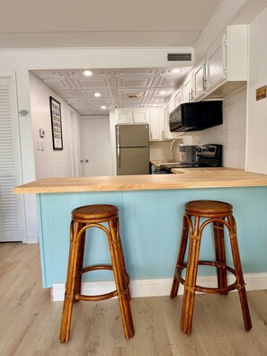 A kitchen with white cabinets, a stainless steel refrigerator, a microwave, and a coffee machine. There are two wooden stools at the island. - Oceanside Condo - Sleeps 6 - Beach Gear - Parking (Ocean City)