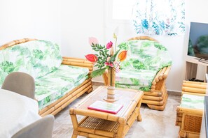 A room with two wicker sofas with green leaf patterns, a small table with a vase of flowers, and a television on a wooden stand. - L'Etoile des îles - À moins d'1 km de la plage (Sainte-Anne)