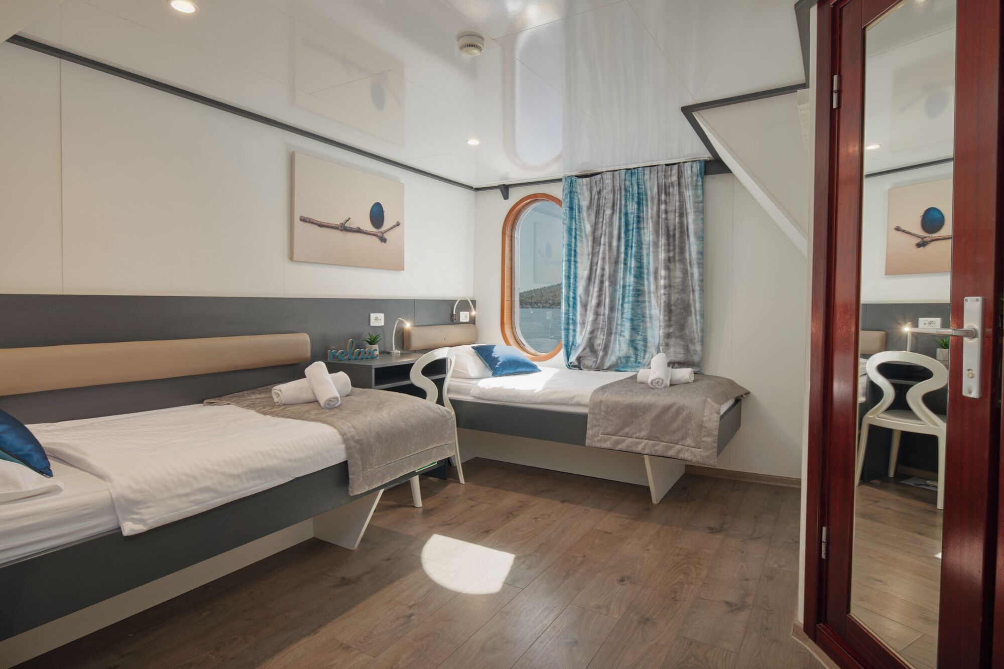 A hotel room with two beds, a desk, a chair, a window with curtains, and a ceiling light.