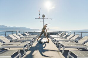 Eine Terrasse mit Liegestühlen, eine Person trainiert mit Kettlebells, im Hintergrund Meer und Berge.