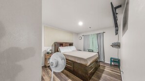 Une chambre d’hôtel comprenant un lit, une table de chevet, une lampe, un ventilateur et une petite table avec une chaise.