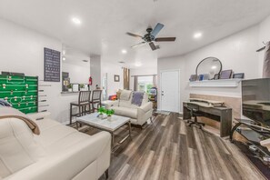 A living room with a sofa, coffee table, and a foosball table. There is a ceiling fan, a TV, and a door leading to another room. - Ile Ayo - BR with Shared Bathroom in a Co-Living Home (1) (Houston)