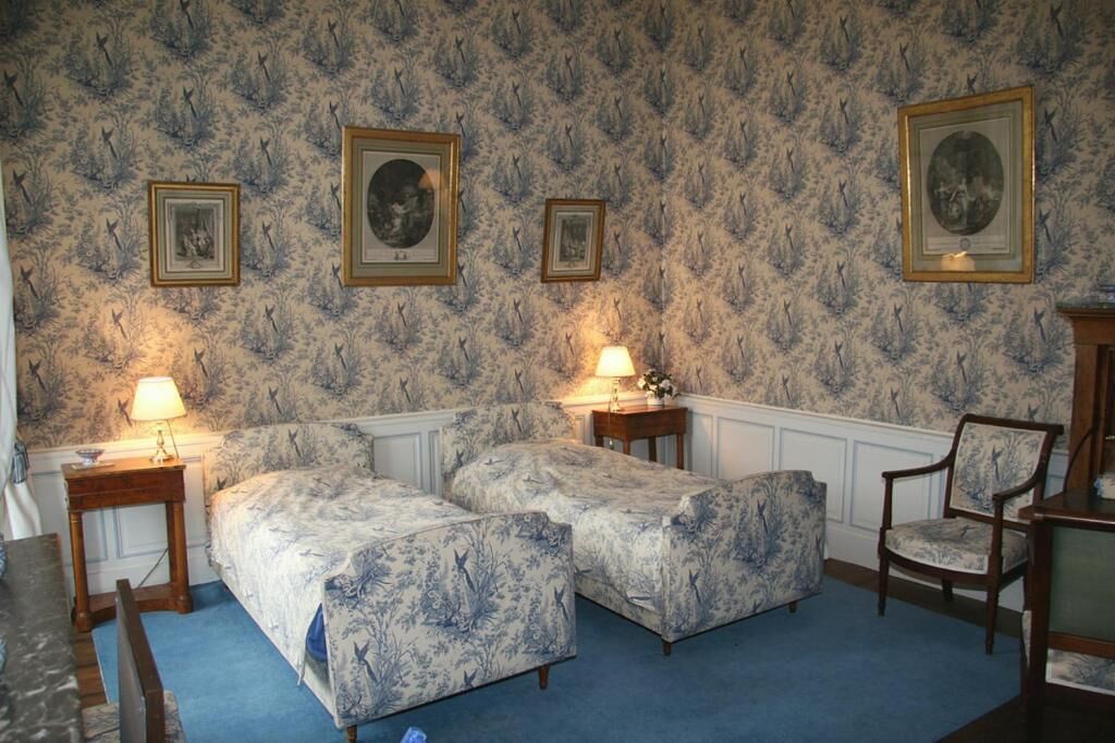 Una habitación con dos camas, una silla, cuadros en la pared y una lámpara sobre una mesita de noche.